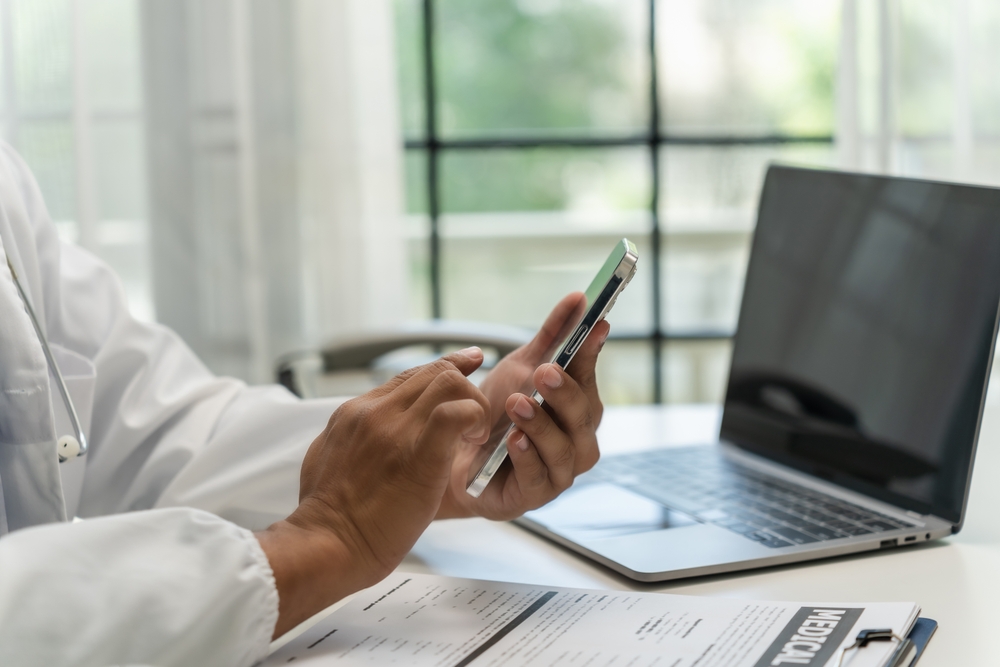 A doctor at a desk is engaged with his phone, possibly checking patient information A doctor at a desk is engaged with his phone, possibly checking patient information