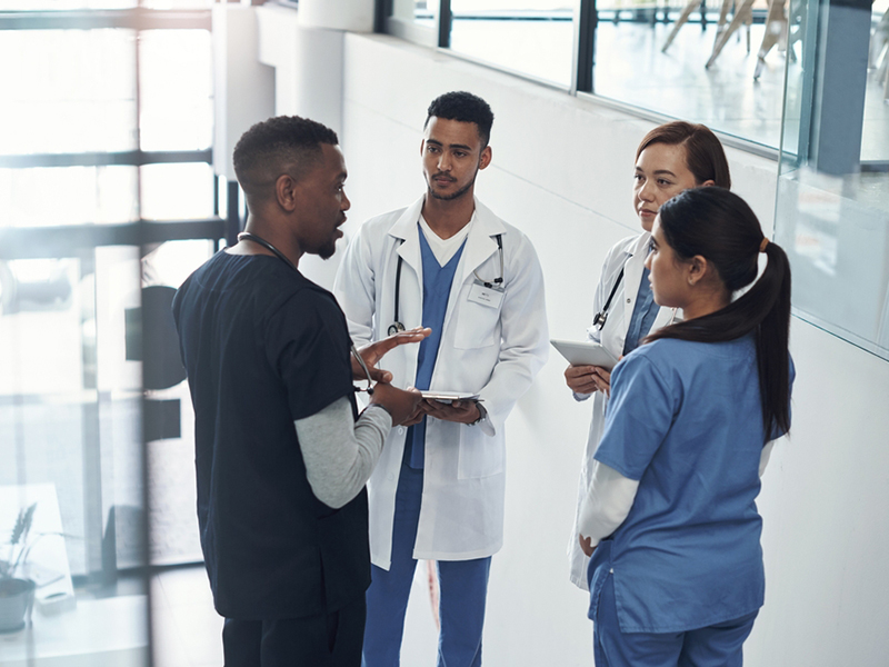 Doctor,,Nurse,And,Group,In,Hospital,With,Team,For,Consult A diverse group of healthcare professionals stands together, wearing scrubs and lab coats, with warm smiles in a bright hospital setting.