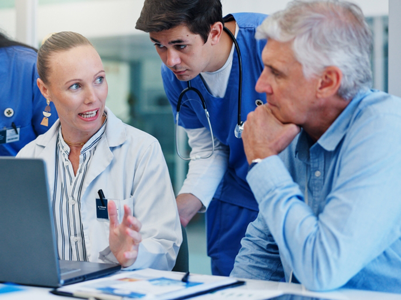 Woman,,Doctors,And,Laptop,With,Group,At,Hospital,With,Discussion An Asian man in a wheelchair sits beside a nurse, who is engaged in conversation and offering care